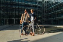 Homem e mulher alegre com bicicleta sorrindo e olhando um para o outro enquanto se comunica fora do prédio de escritórios na rua moderna da cidade — Fotografia de Stock