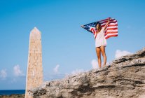 Woman on vacation in casual while shirt standing on big cliff and holding American flag under hand with blue sky and rocked obelisk on background — Stock Photo