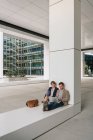 Empresários encantados sorrindo e navegando laptop juntos enquanto sentados do lado de fora do edifício moderno na rua da cidade — Fotografia de Stock