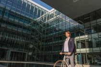 Vista lateral do empresário encantado sorrir e andar de bicicleta no dia ensolarado no centro da cidade moderna — Fotografia de Stock