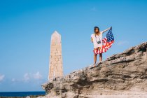 Mujer de vacaciones en casual, mientras que la camisa de pie sobre un gran acantilado y la celebración de la bandera americana bajo la mano con el cielo azul y el obelisco balanceado en el fondo - foto de stock