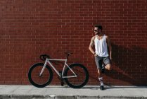 Relajado ciclista masculino moderno en ropa deportiva y gafas de sol descansando junto a la bicicleta mientras está de pie en una pierna y apoyado hacia atrás y el pie en la pared de ladrillo rojo - foto de stock