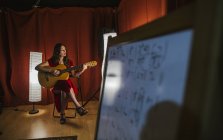 Affascinante donna artistica in abito rosso che suona la canzone alla chitarra sul palco con luce calda in Spagna — Foto stock