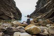 Vue latérale d'une touriste rêveuse en vêtements décontractés assise seule et contemplant sur une plage rocheuse une gorge et une baie tranquille sous un ciel nuageux en Espagne — Photo de stock