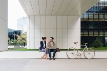 Zufriedene Geschäftsleute lächeln und stöbern gemeinsam am Laptop, während sie vor einem modernen Gebäude in der Nähe von Fahrrädern auf der Stadtstraße sitzen — Stockfoto