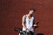 Красивий чоловічий велосипедист у спортивному одязі, використовуючи смартфон, відпочиваючи на велосипеді поруч з червоною цегляною стіною — стокове фото