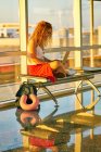 Stilvolle fröhliche Frau tippt auf Laptop, während sie mit gekreuzten Beinen auf Metallbank in Glasflur des Flughafens in Texas sitzt — Stockfoto