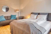 Chambre confortable avec murs en bois blanc et grand lit doux avec canapé gris confortable près du lampadaire en feu — Photo de stock