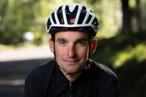 Впевнений дорослий чоловік у велосипедному шоломі посміхається і дивиться на камеру на розмитому тлі парку під час тренувань — стокове фото