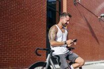 Bonito ciclista masculino em sportswear usando smartphone enquanto descansa na bicicleta ao lado da parede de tijolo vermelho — Fotografia de Stock