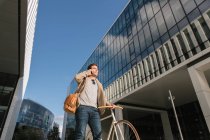 Людина в повсякденному одязі робить телефонний дзвінок, стоячи з велосипедом проти сучасного бізнес-центру висотного підйому зі скляними стінами в центрі міста — стокове фото
