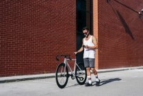 Beau cycliste masculin en vêtements de sport et lunettes de soleil à l'aide d'un smartphone tout en restant debout avec un vélo à côté du mur de briques rouges — Photo de stock