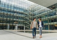 Full body delighted colleagues smiling and looking at each other while walking outside contemporary building after work — Stock Photo