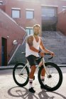 Sportive man wearing white sleeveless shirt and black shorts sitting on bicycle between buildings near stairs on summer sunny day — Stock Photo
