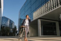 Positive businesswoman with bike smiling and speaking on smartphone while walking outside contemporary building on sunny day on city street — Stock Photo
