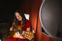 Обаятельная артистка с закрытыми глазами в красном платье исполняет песню, играя на гитаре на сцене с теплым светом в Испании — стоковое фото