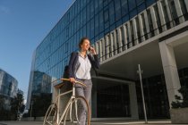 Positive businesswoman with bike smiling and speaking on smartphone while walking outside contemporary building on sunny day on city street — Stock Photo