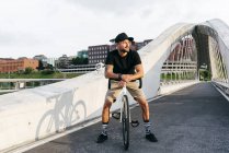 Щасливий дорослий бородатий чоловік у чорній шапці в чорній сорочці та бежевих шортах, що сидить на велосипеді через пішохідний міст у місті, дивлячись на камеру — стокове фото