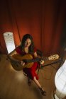 Сверху талантливая женщина в красном платье исполняет песню и играет на гитаре в теплой освещенной сцене рядом с белой лампой — стоковое фото