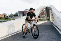 Happy adult bearded man in black cap wearing black shirt and beige shorts sitting resting on bicycle across footbridge in city looking at camera — Stock Photo