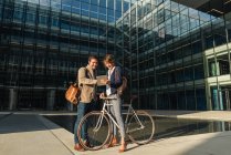 Cheerful man and woman with bicycle smiling and looking at a tablet while communicating outside office building on modern city street — Stock Photo