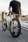 Cropped image of man wearing black shirt and beige shorts standing with white bicycle on bridge with city buildings on background — Stock Photo