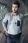 Homme beau à la mode en veste grise élégante et t-shirt décontracté debout et regardant loin avec mur sur fond — Photo de stock