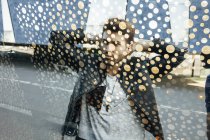 Attraverso la vista vetro decorato di elegante giovane uomo in pelle alla moda giacca nera in piedi con le mani sulla nuca in strada della città — Foto stock