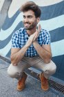 Cheerful young male in casual plaid shirt and sneakers squatting and looking away with painted wall on background — Stock Photo