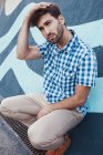 Веселый молодой мужчина в клетчатой рубашке и кроссовках, сидящий на корточках и смотрящий в сторону с раскрашенной стеной на заднем плане — стоковое фото