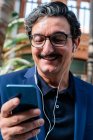 Aged businessman using smartphone with headphones and smiling — Stock Photo