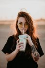 Charming trendy teenage girl in sunglasses and with tattoos enjoying milkshake from glass with straw smiling at camera in sunset — Stock Photo