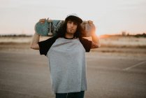 Elegante ragazzo hipster adolescente in possesso di skateboard dietro la testa e guardando la fotocamera sulla strada vuota al tramonto — Foto stock