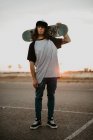 Stylish hipster teenage guy holding skateboard on behind head and looking at camera on empty road in sunset — Stock Photo