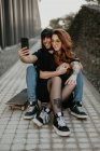 Крута модна пара сидить на дорозі зі скейтбордом і приймає селфі разом з мобільним телефоном у місті — стокове фото