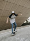 Cool millennial guy in cap balancing while riding skateboard on city street under sloping wall — Stock Photo