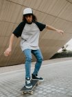 Cool millennial chico en el equilibrio de gorra mientras monta monopatín en la calle de la ciudad bajo la pared inclinada - foto de stock