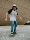 Cool millennial chico en el equilibrio de gorra mientras monta monopatín en la calle de la ciudad bajo la pared inclinada - foto de stock