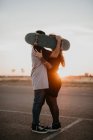 Vista lateral de pareja adolescente romántica abrazando y cubriendo cabezas con monopatín mientras se besa en la espalda iluminada de atardecer en camino rural - foto de stock