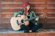 Стильный подросток задумчиво играет на гитаре, сидя на земле со скрещенными ногами и отводя взгляд — стоковое фото