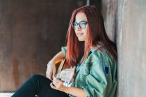 Боковой вид стильного контента подросток, играющий на гитаре, сидя на земле со скрещенными ногами и глядя в камеру — стоковое фото