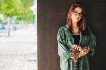 Содержание стильный подросток в очках в темно-зеленой рубашке держа гитару, стоящую рядом коричневая стена, глядя на камеру — стоковое фото