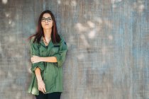 Content stylish teenager in glasses in dark green shirt nearby brown wall looking at camera — Stock Photo