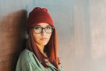Thoughtful stylish teenager in warm hat and glasses in dark green shirt nearby brown wall looking along — Stock Photo