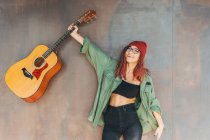 Zufriedener stilvoller Teenager mit Brille in dunkelgrünem Hemd, der eine Gitarre in der Nähe einer braunen Wand hält und in die Kamera blickt — Stockfoto
