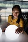 Long haired exited Asian woman watching and touching white illuminating round lamps on table in waiting hall — Stock Photo