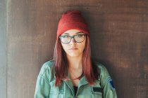 Nachdenklich stilvoller Teenager mit warmem Hut und Brille in dunkelgrünem Hemd neben brauner Wand, der mitschaut — Stockfoto