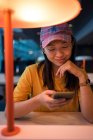 Asian woman in cap surfing on mobile phone sitting at table before schedule board in airport — Stock Photo