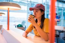 Vue latérale de la femme asiatique en casquette buvant du café à partir de casquette jetable à la table avec éclairage et regardant vers le haut à l'aéroport — Photo de stock