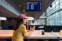 Вид збоку азіатки в кепці серфінг мобільний телефон і пити каву з одноразової кепки за столом в аеропорту — стокове фото